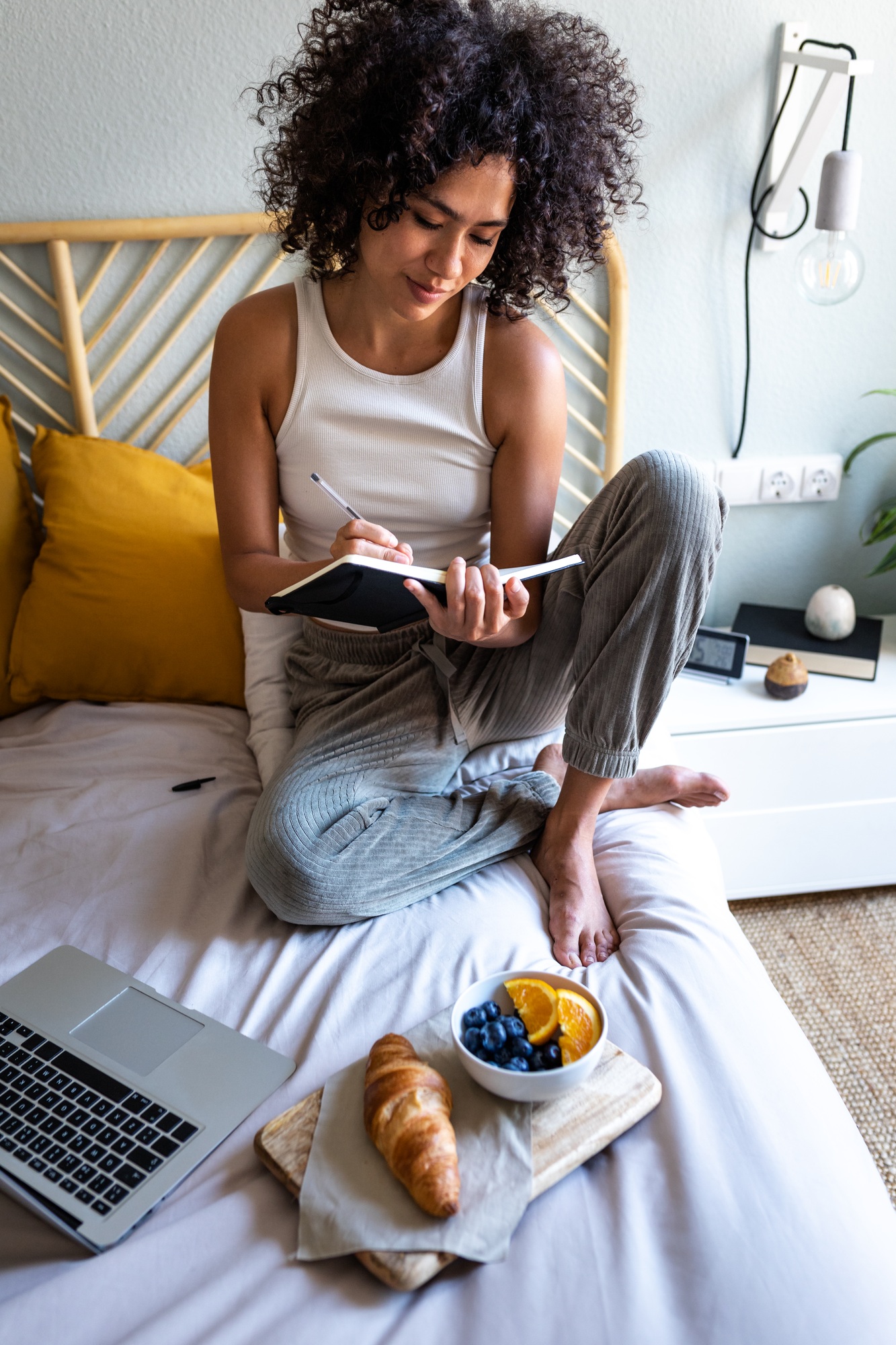 Multiracial woman writing daily affirmations on journal while eating breakfast in bed.Vertical