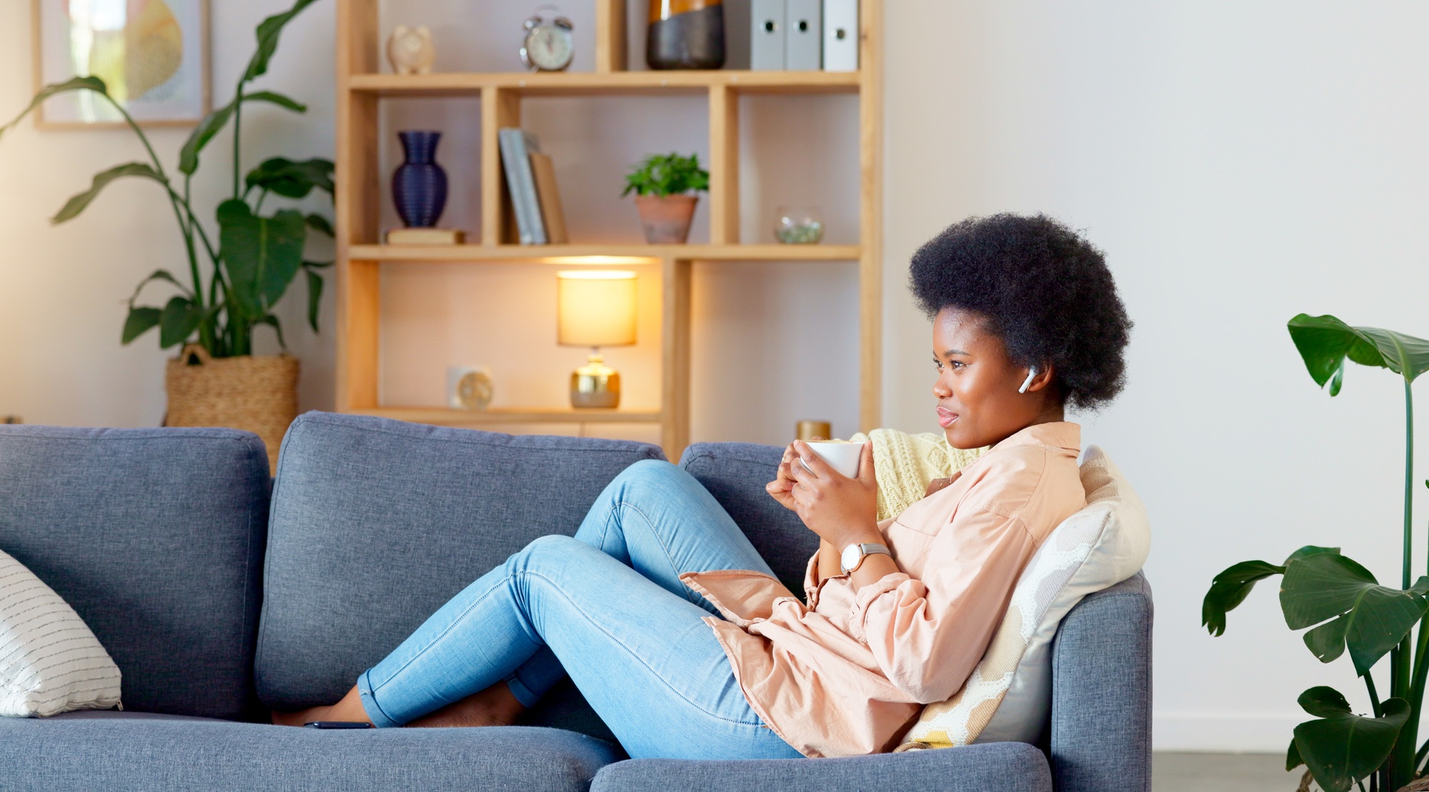 Relaxed Black woman listening to a podcast while drinking coffee and sitting on a couch at home. Ha